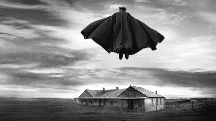 A caped man flies over a desolate farm.