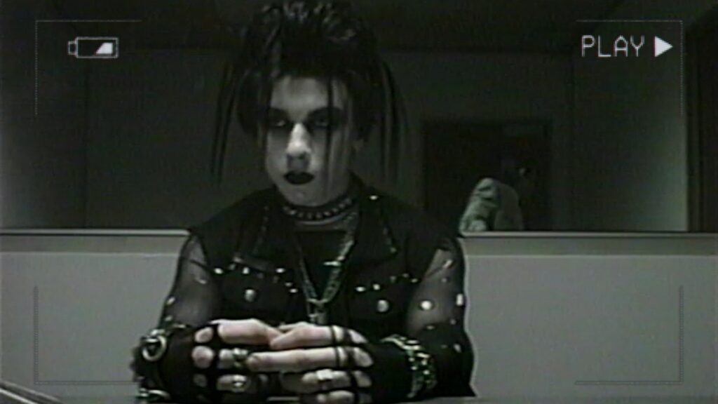 A goth boy waits in an interrogation room. Behind him is a two-way mirror.