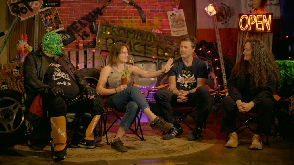 A production still from Disasterpiece which show Fantastico interviewing the Adams family. Lulu sits closest to him holding a bloody mary. She appears to be animatedly telling a story.