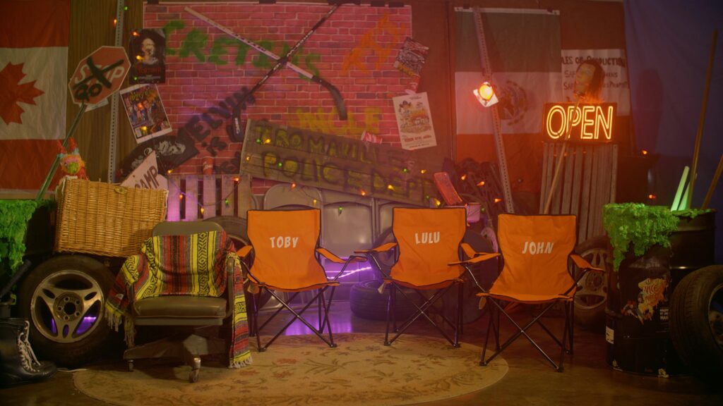 A production still of the Disasterpiece set. It shows Fantasticos empty chair next to three empty orange chairs for the Adams family. The chairs read "Toby," "Lulu," and "John." 