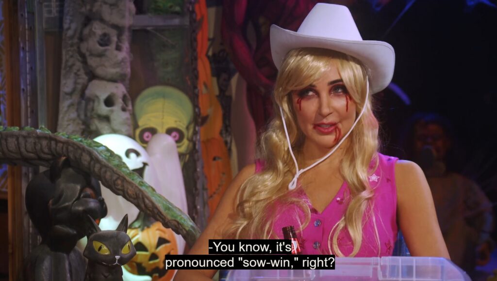 A still image from Joe Bob's Helloween Special on Shudder. It shows Darcy in her Demon Barbie outfit correcting Joe Bob's pronunciation of Samhain. The caption reads "You know, it's pronounced 'sow-win,' right?"