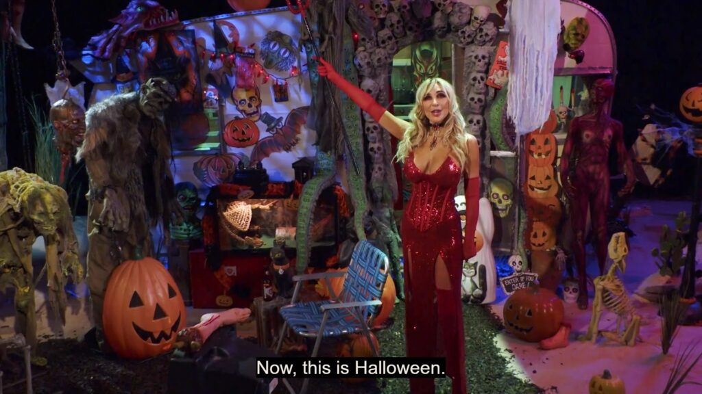 Darcy the Mail Girl stands in a red dress with elbow length red gloves and devil horns. Her arm is outstretched over the set which is covered in Halloween decorations for the Helloween special. The caption reads "Now, this is Halloween." 