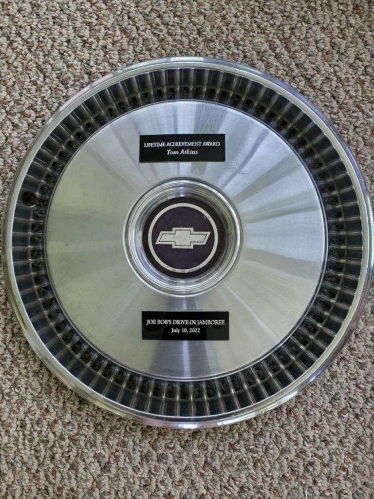 A photograph of the Hubbie award given to Tom Atkins. It shows a Chevrolet hubcap with added inscriptions that read "Lifetime Achievement Award Tom Atkins"
and "Joe Bob's Drive-In Jamboree July 10, 2022."