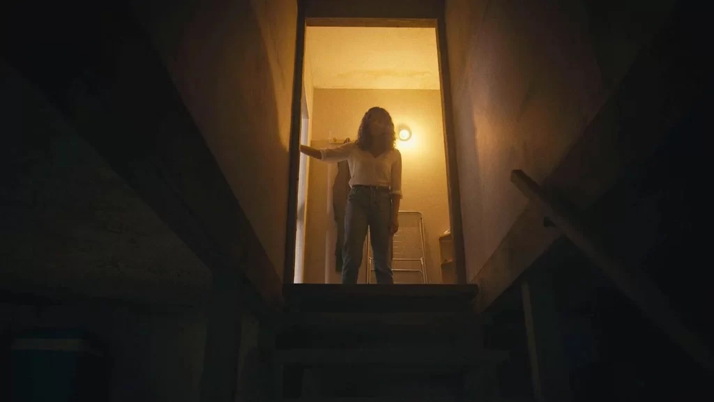 A woman stands above a staircase, the only light behind her as she rests her hand by the door.