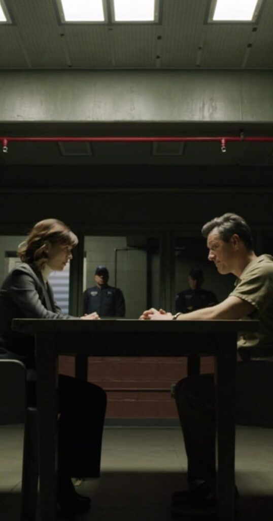 In a prison interrogation room, a woman stares off against a prisoner.