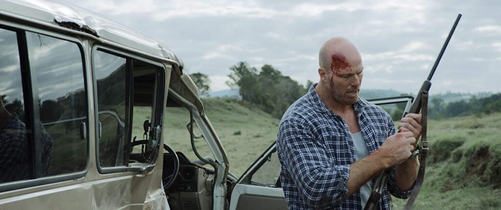 Beside a demolished car, a large man loads his rifle. His head is bleeding, but he remains focused.