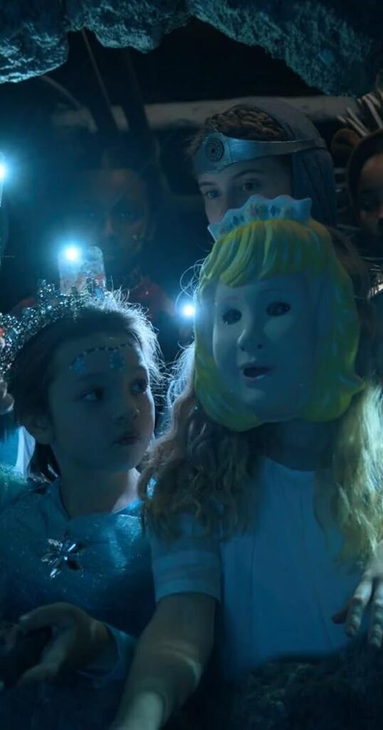 A girl with a princess mask looks ahead. A younger girl looks at the masked girl. Behind them are girls holding cellphone light to see in the night.