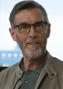 An older man looks amused by something, staring right at the viewer. The background shows a building