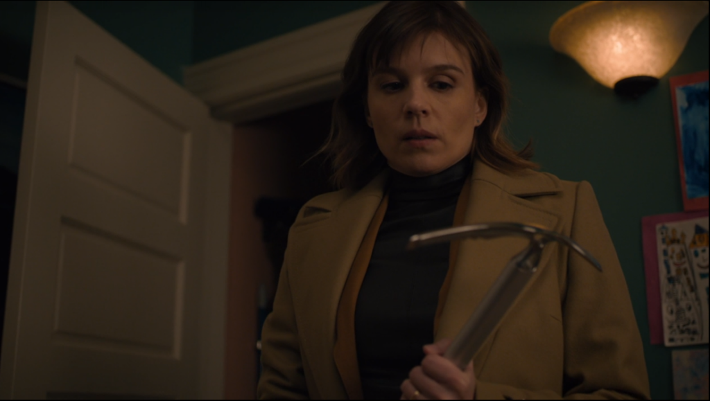 A woman (Kristen) holds an ice axe in her hand. The room seems small, perhaps a closet, with a lamp beside her.