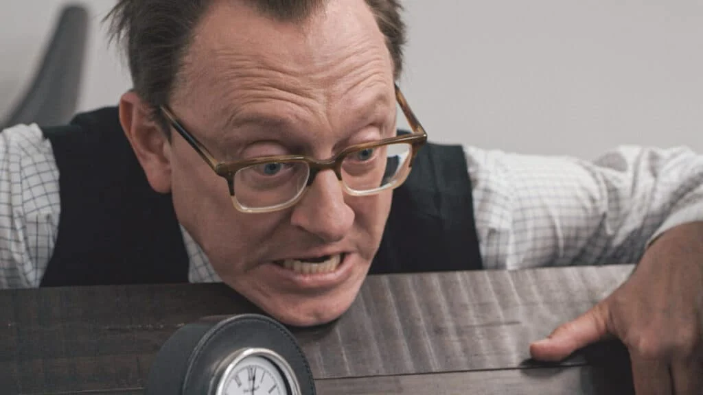 A man wearing glasses clenches a table, looking troubled.