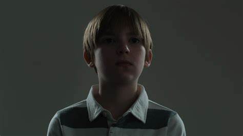 A boy looks onward, shrouded in darkness and over a dark gray background