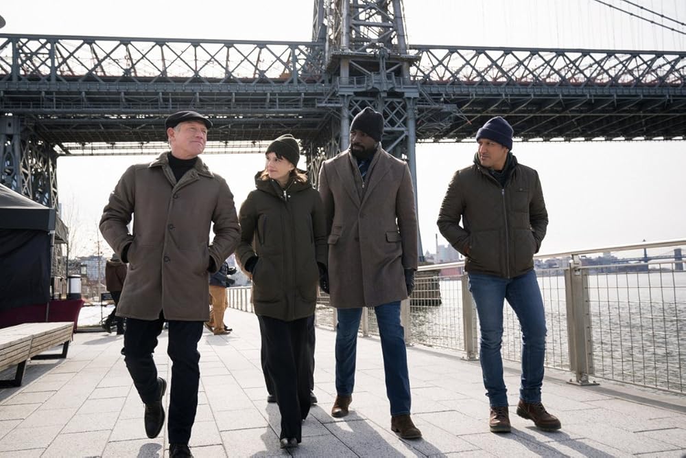 Four people walk beside a river in a city setting. A bridge in the background. An older man talks as the woman and two other men listen.