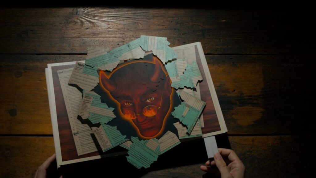 A popup book reveals a red, demonic man with glasses. Underneath the figure is torn 1040 and IRS papers.