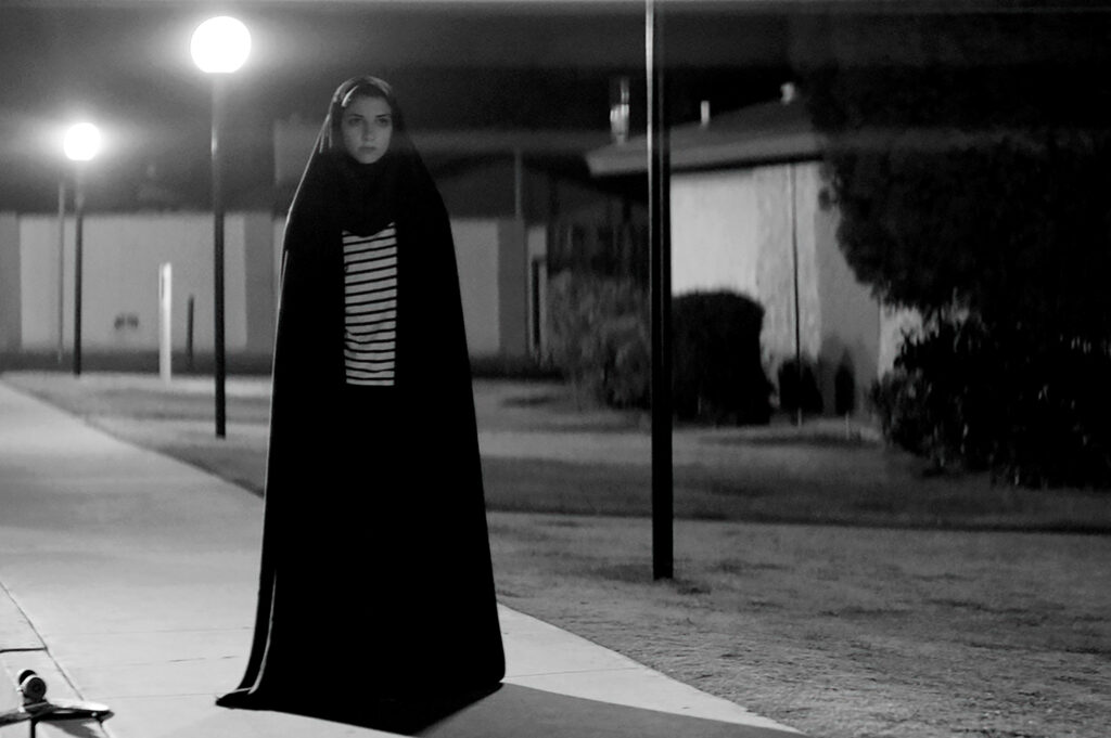 A woman in a hijab and traditional Muslim attire for women stands by lamplight. She glares ahead at someone with a skateboard beside her.