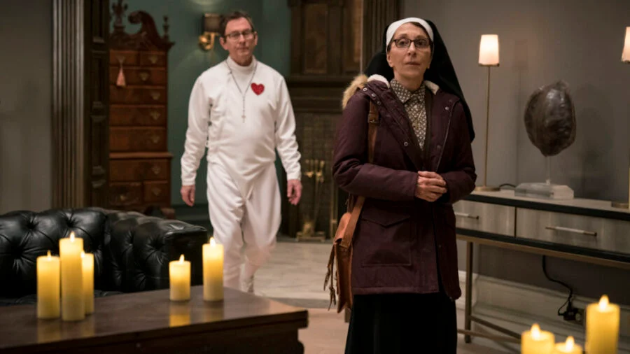 A man dressed in a onesie with a heart on the chest walks behind a nun in a room lit by candles