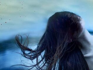 A girl with long hair and her face covered against a blue background with blood splatters.