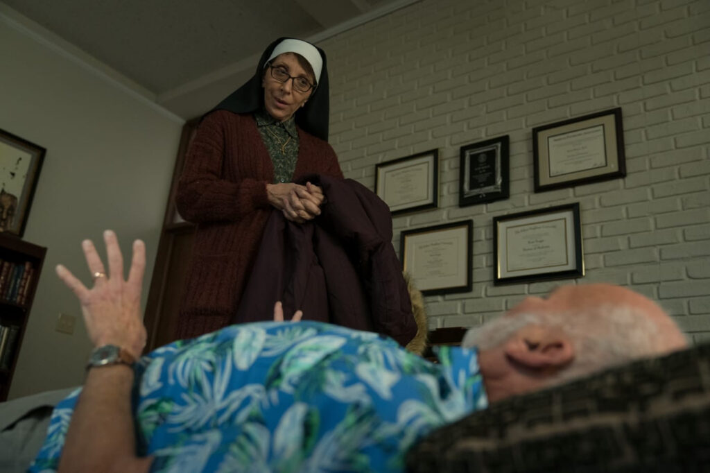 A nun looks down at a therapist who lays on his back. The room suggests a therapists office with certifications lined up on the wall.