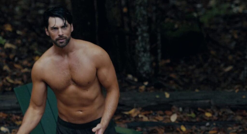 A muscular man looks at the viewer, swaying against the backdrop of a forest