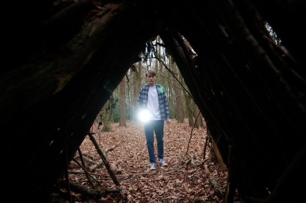 A young man points a flashlight at the viewer. He looks into a triangular opening of a wooden hut. Around him suggests a forest setting.