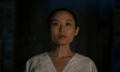 A woman in white is surrounded by darkness, looking ahead with a poker face.