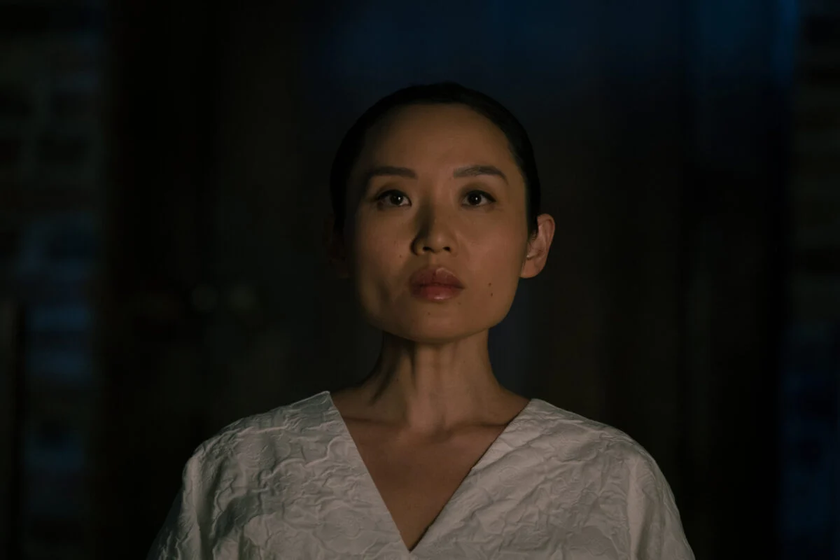 A woman in white is surrounded by darkness, looking ahead with a poker face.