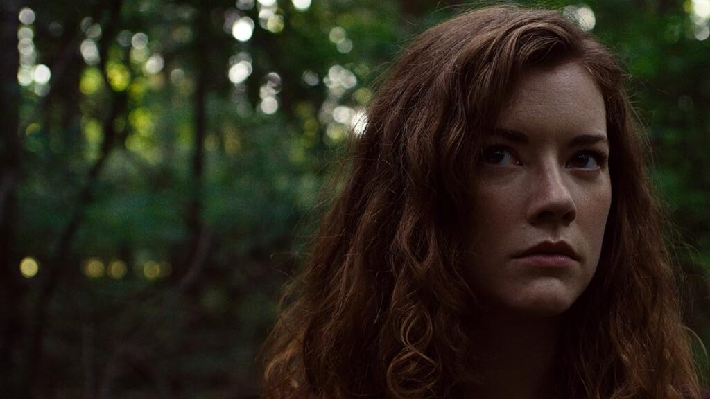 A woman stares in the distance, focused on the situation. Behind her is a lush forest.