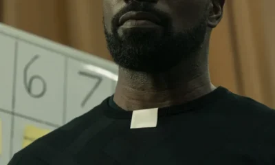 A man looks in confusion. He wears a black shirt with a white paper acting as the Roman collar of a priest.