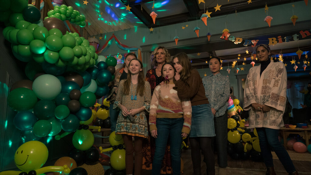 A party setting with dominant green balloons. Several people stand to celebrate someone unseen.