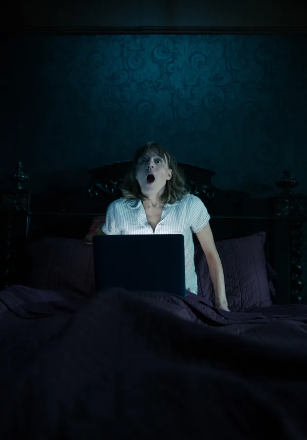 A woman emerges from her bed, looking shocked. The laptop on her lap is the only source of light.